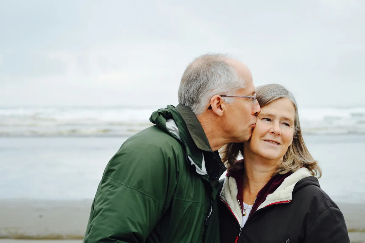 An older couple standing close together by the sea, wind in their hair