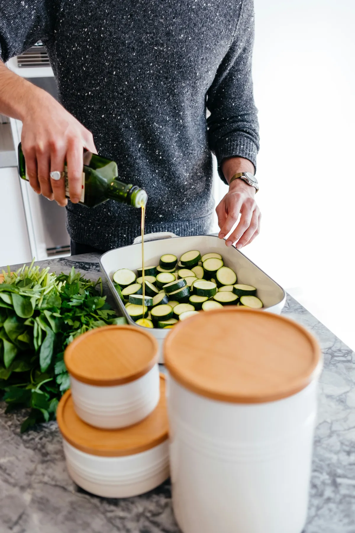 Hands pouring olive oil over a tray of sliced courgettes on a kitchen counter