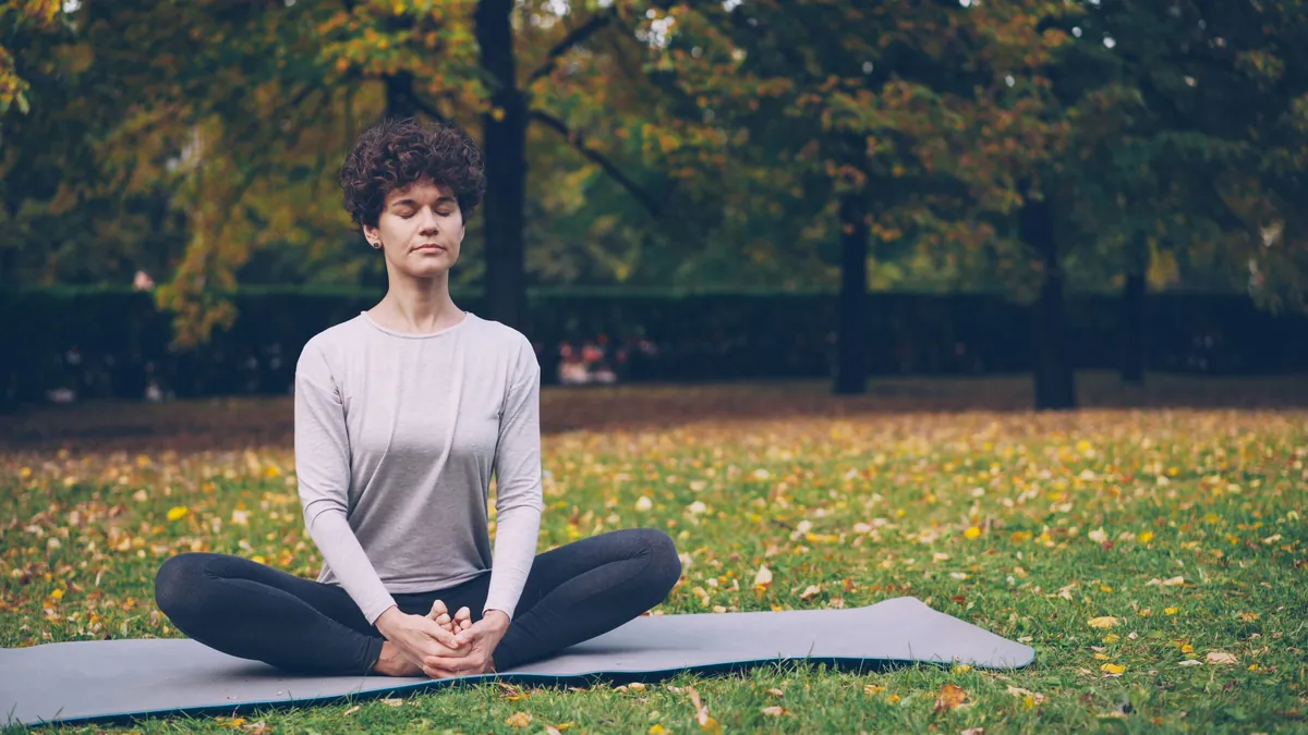 A woman sitting cross-legged on a mat in a park in autumn, eyes closed, leaves on the grass around her